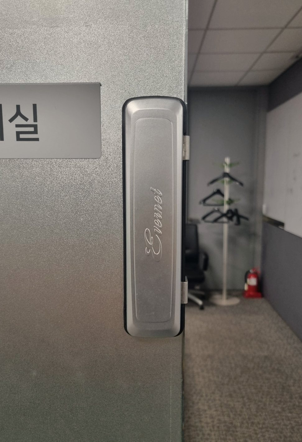 일산서구 대화동 출장 — 반투명 유리문 가장자리에 붙은 길쭉한 은빛 도어락, 문 너머의 사무실 옷걸이와 소화기